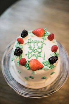 a birthday cake with white frosting and berries on top is sitting on a glass plate