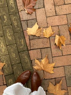 a person's feet in white pants and brown shoes on a brick walkway with leaves scattered around them