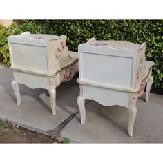 two white nightstands with pink flowers painted on them sitting in front of a bush