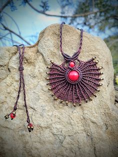 a purple necklace with red beads on it sitting on top of a rock next to a tree