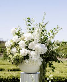 white flowers are in a large vase on a pedestal outside with hedges and bushes behind it