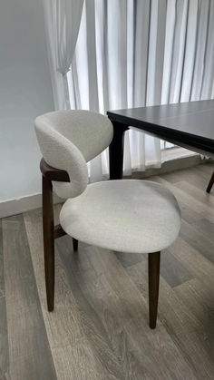 A modern dining chair with a unique design, featuring a rounded backrest and a seat upholstered in a textured light beige fabric. The chair stands on slender, dark wooden legs, contrasting with the light flooring. Behind it, a sleek black dining table edge is visible, and the scene is set against a backdrop of flowing sheer white curtains. The overall atmosphere is minimalist and elegant, suggesting a blend of comfort and contemporary style. Chic Dining Room, Modern Dining Chairs, Textured Fabric, Dining Area