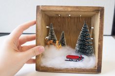 someone is holding up a miniature christmas scene in a box with trees and a red truck