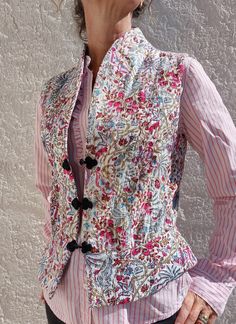 a woman wearing a pink and white shirt with a floral vest on her shoulders, standing in front of a gray wall