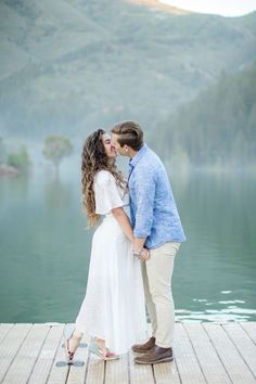 a man and woman kissing on a dock by the water with mountains in the background