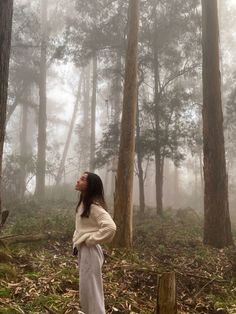 a woman standing in the middle of a forest looking up into the foggy sky