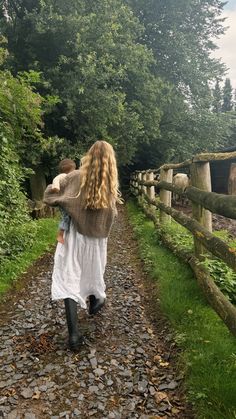 a woman walking down a dirt road next to a lush green forest filled with trees