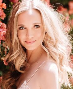 a woman with long blonde hair and blue eyes posing in front of some pink flowers