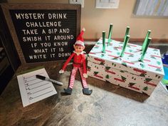 an elf is sitting next to a box with writing on it and a sign that says, mystery drink challenge take a sip and finish it around if you know what it is write it down