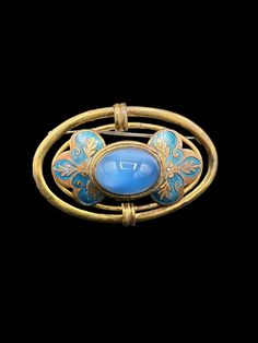 This vintage faux moonstone brooch is stunning. The large oval brass setting, adorned with blue enamel floral decorations, encircles a large sky-blue glass cabochon reminiscent of a moonstone. Its manufacturing date is likely in the 1940s. The glass cabochon could be Czech, but I'm not positive. With its considerable size, the brooch could be a striking scarf pin. While it does show signs of its age with some blemishes to the brass, it still maintains its beauty. To ensure you have a comprehensi Luxury Oval Cabochon Brooch, Blue Oval Brooch Jewelry, Oval Blue Brooch Jewelry, Oval Blue Brooch, Vintage Oval Brooch With Cabochon, Vintage Oval Cabochon Brooch, Blue Oval Brooch, Antique Blue Enamel Brooches, Vintage Oval Brooches For Collectors