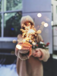 a woman holding a sparkler in her hand with christmas lights on the window sill behind her