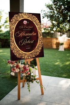 a sign with flowers on it in front of some grass and trees at a wedding