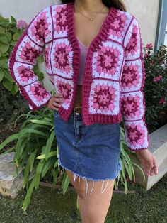 a woman wearing a pink and white crochet jacket standing in front of some bushes