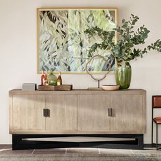 a large painting hangs on the wall above a wooden cabinet with two vases filled with plants