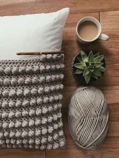 a crocheted pillow and yarn next to a cup of coffee on a wooden floor