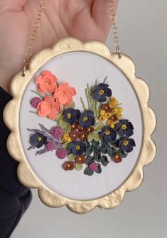 a hand holding a small embroidered flower decoration on a white background with gold trimmings