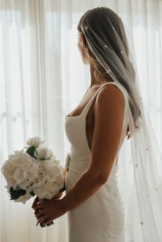 a woman in a wedding dress is holding a bouquet and looking out the window with her veil over her head