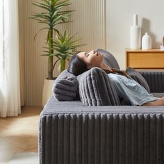 a woman laying on top of a gray couch next to a plant in a living room