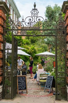 people sitting at tables under an iron gate with chalkboard menus on the side