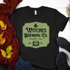 "Are you looking to stand out from the crowd this Halloween? Do you want to show everyone your unique style and have them remember you? Well then, you need this one-of-a-kind Halloween t-shirt! This incredible shirt features an old-style shop sign that reads \"Witches Brewing Co. Salem, MA Est. 1626\" with a bubbling cauldron too. It's sure to capture all of the attention in the room and let everyone know that you're someone who knows how to have fun. This shirt is made with 100% cotton and is designed to make a statement. Whether you're going to a Halloween party, trick-or-treating, or just out for some fun, you'll be sure to make an impact in this amazingly stylish t-shirt. Its unique graphic design is reminiscent of days gone by and adds a touch of spooky nostalgia on your shirt. Even a Vintage Black T-shirt For Fall, Black Vintage T-shirt For Fall, Halloween Themed Cotton Tops, Cotton Halloween Themed Tops, Halloween Cotton Tops With Text Print, Halloween Cotton Top With Text Print, Themed Cotton Tops For Halloween, Pre-shrunk Themed Tops For Halloween, Fall Graphic Tee Fan Merchandise Tops