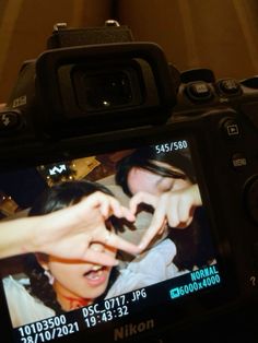 a person making a heart shape with their hand on a camera lens, in front of a woman's face