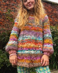 a woman standing in front of a brick wall wearing colorful knitted sweater and pants