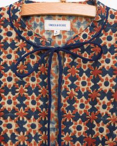 an orange and blue flowered shirt with a bow tie at the neck, hanging on a wooden hanger