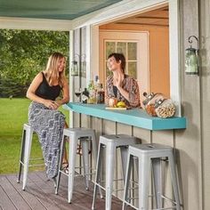 two women sitting at a bar on the back deck of a house, talking to each other