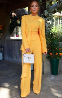 a woman standing in front of a tree holding a white purse and wearing yellow pants