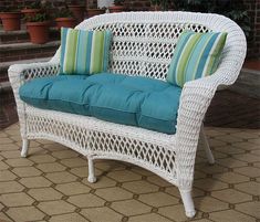 a white wicker couch with blue and green pillows sitting on a patio floor next to potted plants