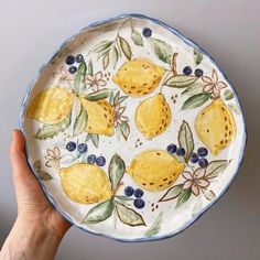 a hand holding a plate with lemons and blueberries painted on the side, in front of a gray wall