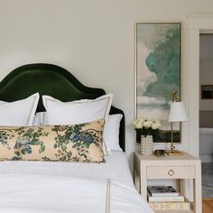 An emerald green velvet headboard accents a bed dressed in white and tan bedding topped with a beige lumbar pillow accented with a botanical design.