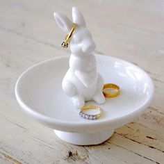 a small white rabbit figurine sitting on top of a plate with two wedding rings