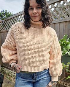 a woman standing in front of a wooden fence wearing a sweater and jeans with tattoos on her arms