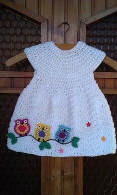 a white crocheted dress with owls and flowers on the front is hanging from a wooden hanger