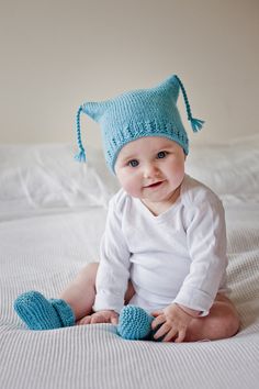 a baby sitting on a bed wearing a blue knitted cat hat and mittens