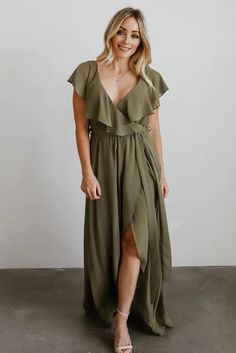 a woman wearing a green dress and sandals standing in front of a white wall with her hands on her hips