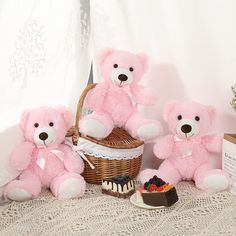 three pink teddy bears sitting in front of a basket with cake and strawberries on the table