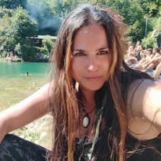 a woman with long hair sitting in front of a body of water and people standing around her