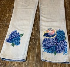 two pairs of white jeans with blue flowers and a bird embroidered on the side, sitting on a wooden floor