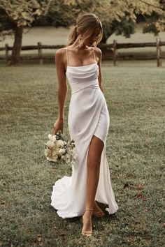 a woman in a white dress holding a bouquet and walking through the grass with her hand on her hip