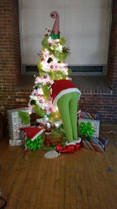 a woman in green pants and santa hat standing next to a christmas tree with presents under it