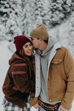 a man and woman standing next to each other in front of snow covered trees wearing hats