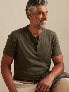 a man with grey hair and beard sitting on a stool smiling at the camera while wearing a green shirt