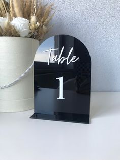 a black table number holder next to a white vase with dried flowers and a ribbon