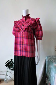 Cute French Vintage short sleeve blouse from the 1980s. Pie crust collar, straight cut. Beautiful Madras check pattern with pink, red, blue and yellow tones. Ruffle bib and yoke. Short puff sleeves with buttoned cuffs. Side vents. Made in France! BRAND: Daniel Duval Paris ERA: 1980s COLOR: Pink, red, yellow, purple, blue FABRIC: 65% polyester, 35% cotton SIZE: Vintage size 40 (FR), fits best size M (or size S for an oversized fit) - for reference please check the measurements below MEASUREMENTS: Plaid Short Sleeve Blouse For Work, Short Sleeve Plaid Tops With Ruffles, Plaid Short Sleeve Tops With Ruffles, Retro Short Sleeve Plaid Blouse, Retro Plaid Short Sleeve Blouse, Vintage Short-sleeve Blouse With Ruffles, Check Blouse, Checked Blouse, Vintage Short