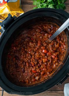 a crock pot filled with chili and beans on top of a wooden table next to chips