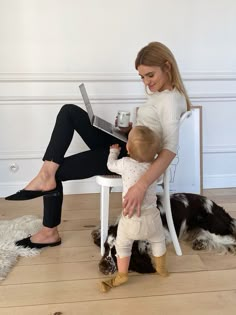 a woman sitting on a chair holding a baby while reading a book to a dog