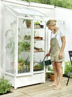 a woman is watering plants in a small white greenhouse on a wooden deck next to a table and chairs