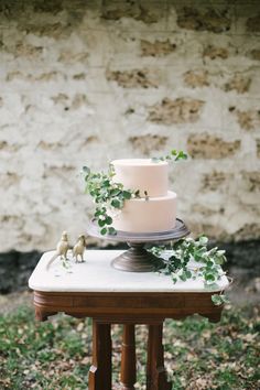 a white wedding cake with greenery on top sitting on a table in front of a brick wall